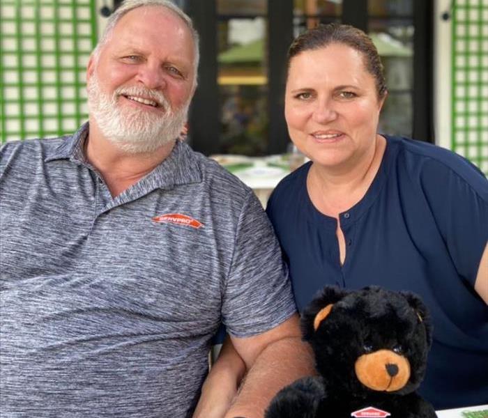 Man and woman sitting in front of restaurant with a teddy bear on the table. 