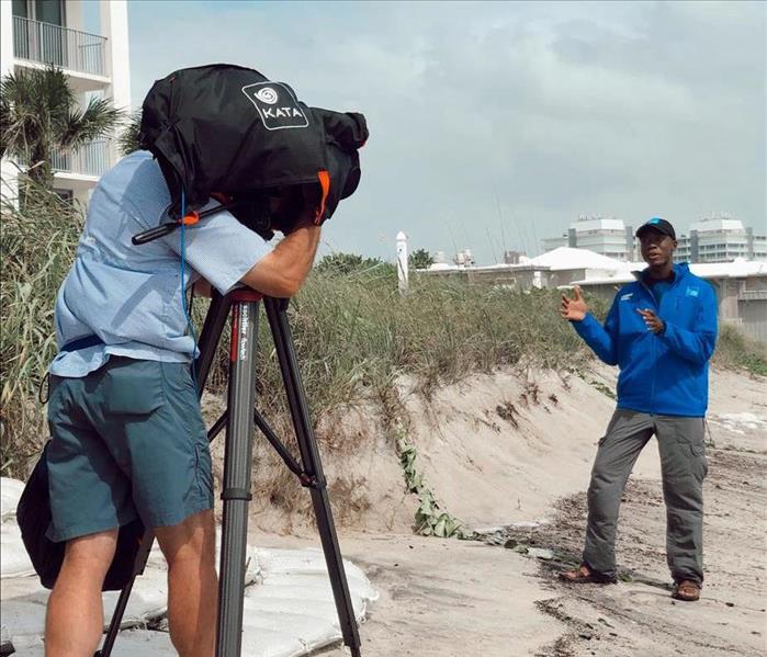 weather man on beach being recorded by a man with a big camera 