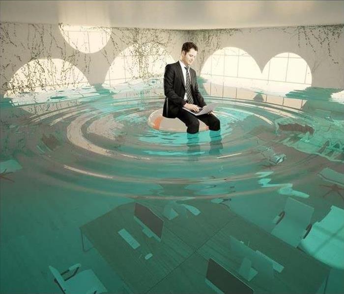Stock photo of a man sitting on a pool float, working on his laptop, inside a flooded office.