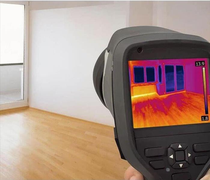 Infrared device pointed towards wall of empty room with white walls and wood flooring to detect leaks behind walls. 