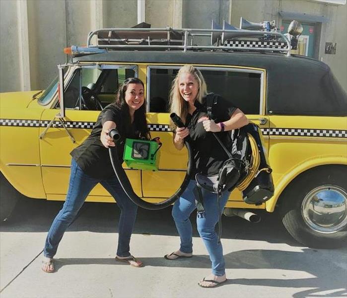 Two women in front of taxi cab posing with deodorizing equipment. 
