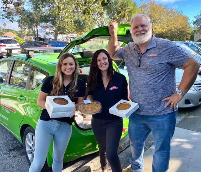 Two women holding pie and a man standing in front of a SERVPRO Vehicle